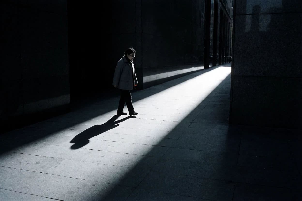 Black and white street photograph of a figure crossing a bright band of sunlight between dark buildings, generated with Krea 2 Medium