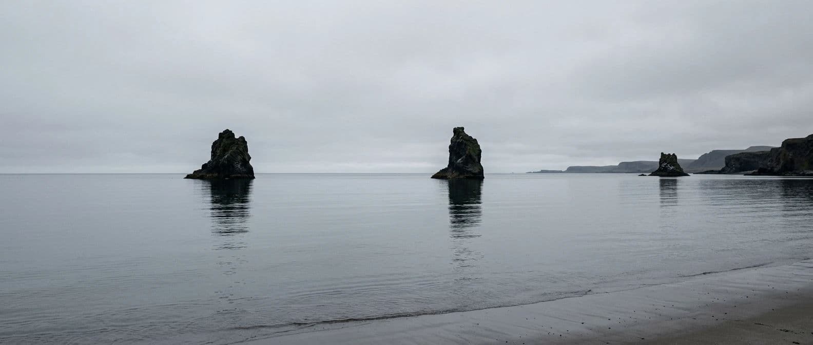 Wide panoramic coastal photograph of three sea stacks spaced across still water under flat grey cloud, generated with Nano Banana 2