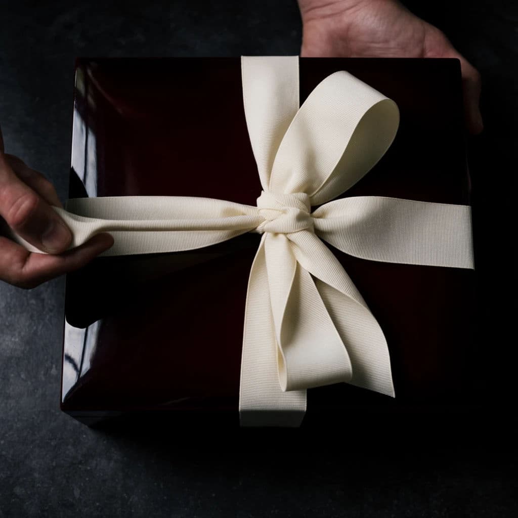 Square photograph of a hand pulling a grosgrain ribbon loose on a deep lacquer box, generated with Krea 2 Large